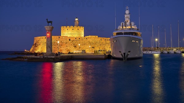 An illuminated castle and a luxury yacht lie by the sea at night with glowing reflections in the water, night shot, deer statue, Fort Agios Nikolaos, Mandraki Oat, Rhodes Town, Rhodes, Dodecanese, Greek Islands, Greece, Europe
