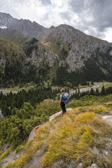 A female hiker stands on a grassy slope against an impressive mountain backdrop under a cloudy sky, Chong Kyzyl Suu Valley, Terskey Ala Too, Tien-Shan Mountains, Kyrgyzstan, Asia