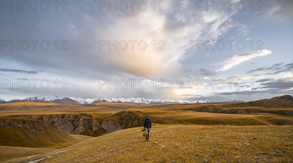 Tourist with camera on plateau with yellow grass, Sary Jaz mountain valley, high glaciated mountain peaks of the Tien Shan in the background, autumnal mountain landscape, Tien Shan, Kyrgyzstan, Asia