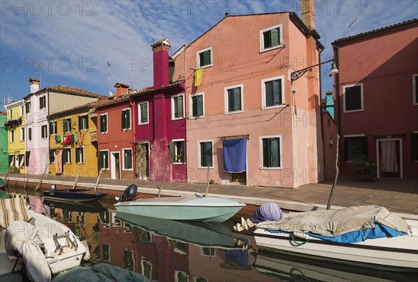 Moored boats on canal lined with pink, red, orange, yellow and green stucco houses decorated with curtains over entrance doors plus washed clothes on clothesline, Burano Island, Venetian Lagoon, Venice, Veneto, Italy, Europe