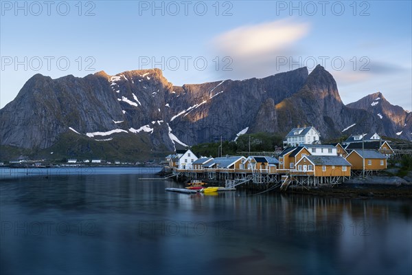The village of Sakrisoy with its typical yellow or ochre-coloured wooden houses on wooden stilts (rorbuer) by the sea. At night during the midnight sun, good weather. Early summer. Long exposure, a blurred cloud in the blue sky. Sakrisoy, Moskenesoya, Lofoten, Norway, Europe