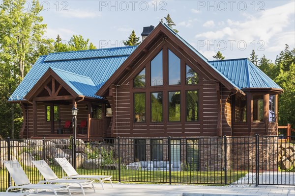 Brown stained milled Eastern white pine timber and flat log profile home facade with stone cladding on walk-out lower level and blue standing-seam sheet metal roof in summer, Quebec, Canada, North America