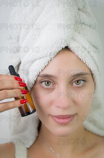 Happy young woman with towel on head holding amber glass bottle of skin serum. Beautiful young Brazilian woman moisturizes her skin with serum isolated against white background