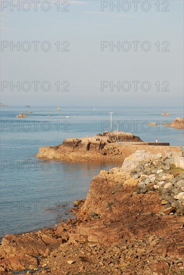 Coastline with rocks, clear calm water, a small lighthouse, and a blue sky reflecting serenity