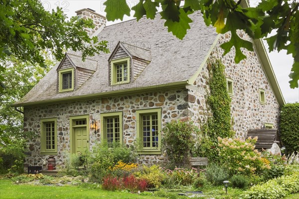 Old circa 1735 Canadiana fieldstone house facade with lime green trim and cedar wood shingles roof and landscaped front yard in late summer, Quebec, Canada, North America