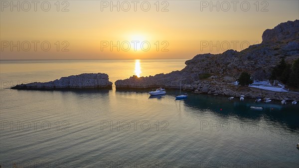 Golden glowing sunset over the calm sea next to rocky cliffs, Paulus Bay, below the Acropolis of Lindos, Lindos, Rhodes, Dodecanese, Greek Islands, Greece, Europe