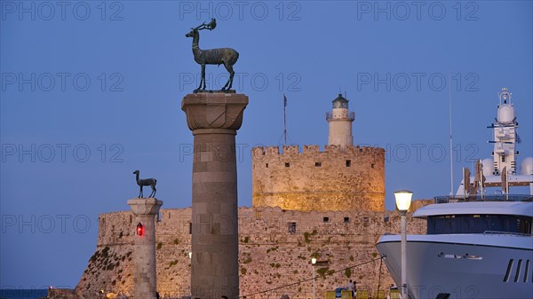 A deer sculpture on a pillar in front of a castle and lighthouse next to a luxury yacht at dusk, Deer statue, Deer statue, Fort Agios Nikolaos, Mandraki harbour, European roe deer, Rhodes town, Rhodes, Dodecanese, Greek Islands, Greece, Europe