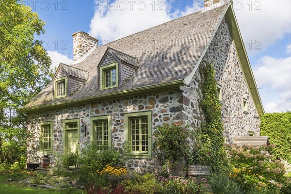 Old circa 1735 Canadiana fieldstone house facade with lime green trim and cedar wood shingles roof and landscaped front yard in late summer, Quebec, Canada, North America