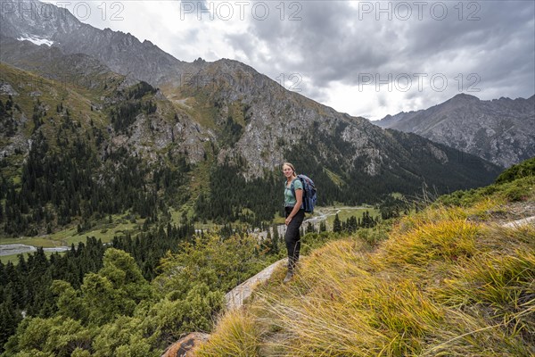 A female hiker stands on a grassy slope against an impressive mountain backdrop under a cloudy sky, Chong Kyzyl Suu Valley, Terskey Ala Too, Tien-Shan Mountains, Kyrgyzstan, Asia