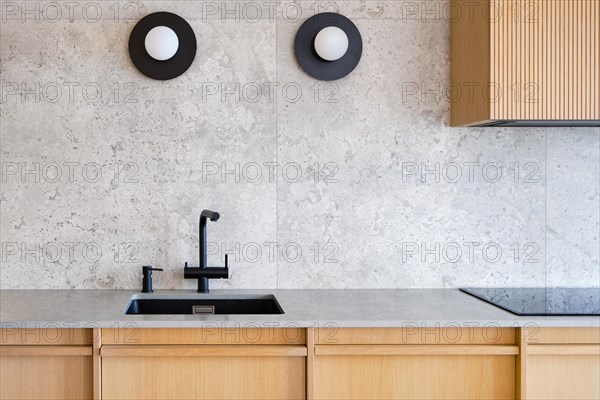 Contemporary, minimalist kitchen interior featuring a sleek black sink and faucet set against a textured grey wall, complemented by two modern circular lights and wooden cabinetry