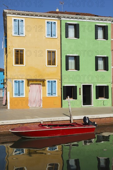 Moored red boat on canal lined with orange and green stucco houses decorated with striped curtains over entrance doors, Burano Island, Venetian Lagoon, Venice, Veneto, Italy, Europe