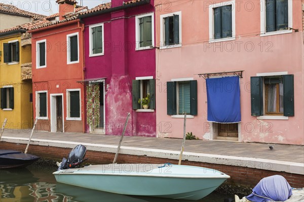 Moored boats in canal lined with pink, purple, red and yellow stucco houses decorated with curtains over entrance doors, Burano Island, Venetian Lagoon, Venice, Veneto, Italy, Europe