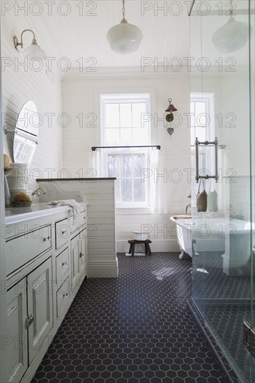 White antique style finish vanity with quartz countertop and two sunken sinks, freestanding claw foot bathtub, glass shower stall in kid's upstairs floor bathroom with black honeycomb ceramic tile floor inside country style home, Quebec, Canada, North America