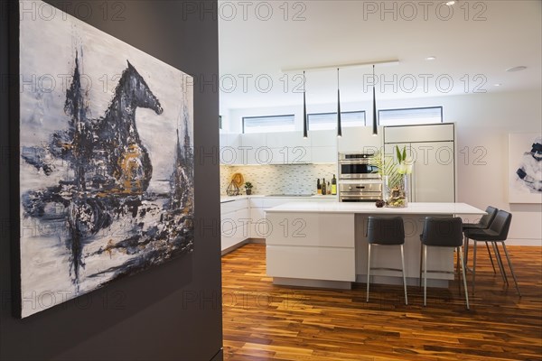 Decorative acrylic painting of mystic horse and castle by Liza Castro-Interior Designer on dark living room wall plus white high-gloss lacquered cabinets and island with grey leather bar stools and white quartz countertops in kitchen with acacia wood floorboards inside contemporary home, Quebec, Canada. Artwork by Liza Castro