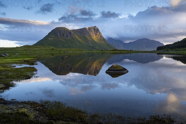 Landscape on the Lofoten Islands. The mountain Offersoykammen and the Beech Vagspollen. Mount Stornappstinden on Flakstadoya in the background. The mountains are reflected in the water. At night during the midnight sun. A few clouds in the sky. The night sun shines on the mountain peak. Early summer. Vestvagoya, Lofoten, Norway, Europe