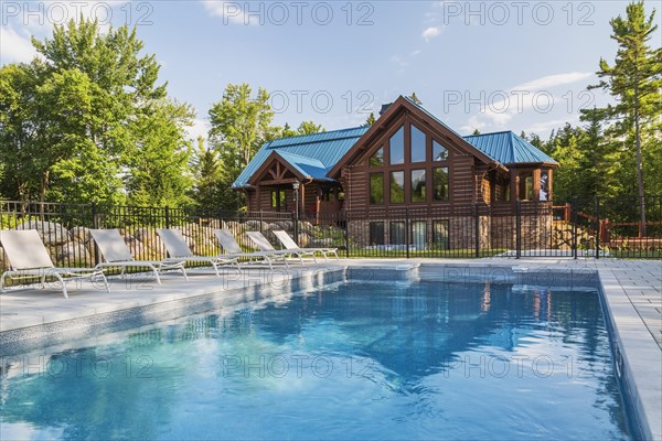 In-ground salt water swimming pool and brown stained milled Eastern white pine timber and flat log profile home facade with stone cladding on walk-out lower level and blue standing-seam sheet metal roof, Quebec, Canada, North America