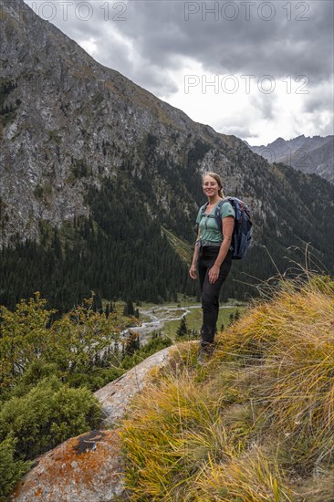 A female hiker stands on a grassy slope against an impressive mountain backdrop under a cloudy sky, Chong Kyzyl Suu Valley, Terskey Ala Too, Tien-Shan Mountains, Kyrgyzstan, Asia