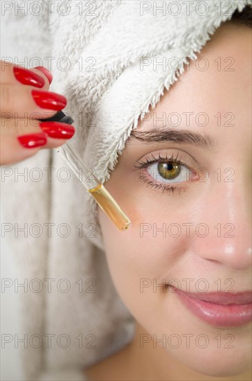 Happy young woman applying serum on her face with her eyes open. Beautiful young Brazilian woman moisturizes her skin with serum isolated against white background