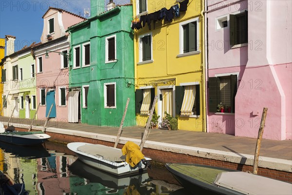 Moored boats on canal lined with pink, yellow and green stucco houses decorated with striped curtains over entrance doors and windows plus clothes on clothesline, Burano Island, Venetian Lagoon, Venice, Veneto, Italy, Europe