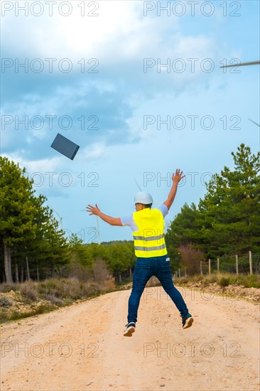 Vertical photo of the rear view of a male adult caucasian happy engineer jumping celebrating in a path next to wind turbine