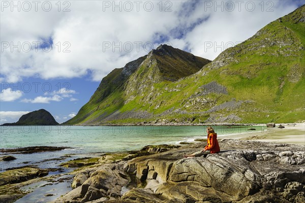 Landscape with sea at the sandy beach of Haukland (Hauklandstranda) with the mountain Veggen. A woman sits barefoot on a rock and looks out to sea. Shot during the day with blue sky. Early summer. Haukland Beach, Haukland, Vestvagoya, Lofoten, Norway, Europe