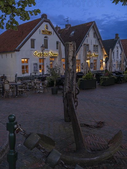 Ship's anchor on a wooden pile in front of restaurants at Greetsiel harbour at night, Greetsiel, Krummhoern, East Frisia, Lower Saxony, Germany, Europe