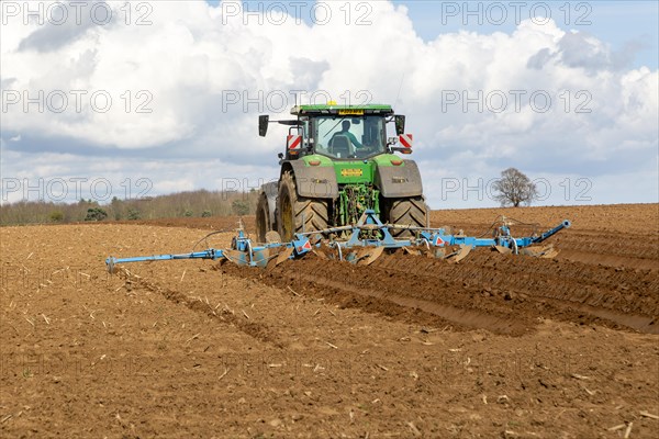 Green John Deere tractor ploughing deep furrows to prepare soil for potato crop, Ramsholt, Suffolk, England, UK