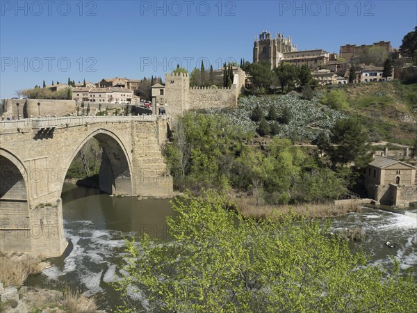 Stone bridge over the river, green hills with historical buildings and cathedral, clear blue sky, historical stone bridge with an old tower and an old city in the background over a wild river, Toledo, Spain, Europe