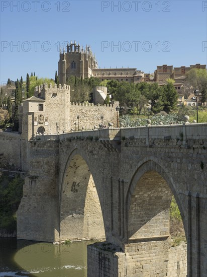 Stone bridge with arches over a river, in the background a church and cityscape, sunny weather, historic stone bridge with an old tower and an old town in the background over a wild river, Toledo, Spain, Europe