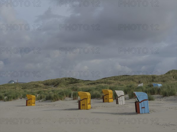 Beach chairs scattered on the beach under a cloudy sky, surrounded by gentle dunes, colourful beach chairs on the beach and in the dunes under an overcast sky, red wooden boat and yellow beach chair, Juist, Germany, Europe