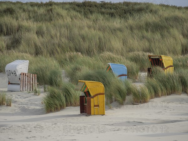 Several beach chairs in different colours, surrounded by high grasses in a quiet dune landscape, colourful beach chairs on the beach and in the dunes under cloudy skies, red wooden boat and yellow beach chair, Juist, Germany, Europe