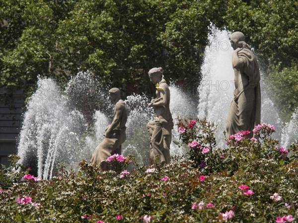 Three statues in a fountain surrounded by blooming flowers and bubbling water fountains, fountain with figures in front of a historic building, barcelona, spain