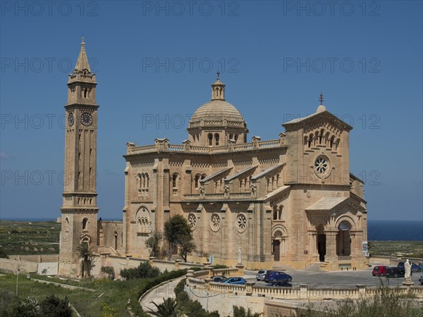 Neo-Romanesque basilica made of light-coloured stones with a high bell tower and some cars in the foreground, isolated church on an island with the blue sea in the background, gozo, malta