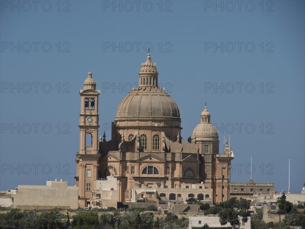 A large baroque basilica with a dome and detailed sandstone architecture set against a blue sky, solitary church on an island with the blue sea in the background, gozo, malta