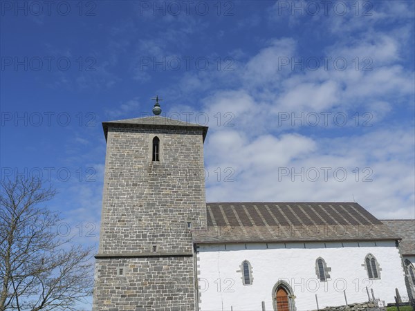 Historic church with stone tower and blue sky, some clouds in the background, old stone church and many gravestones at the water with ships in the background, haugesund, norway