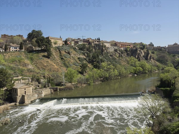 A city view on a hill with buildings, a river with a waterfall and green areas under a clear sky, historic stone bridge with an old tower and an old city in the background over a wild river, Toledo, Spain, Europe