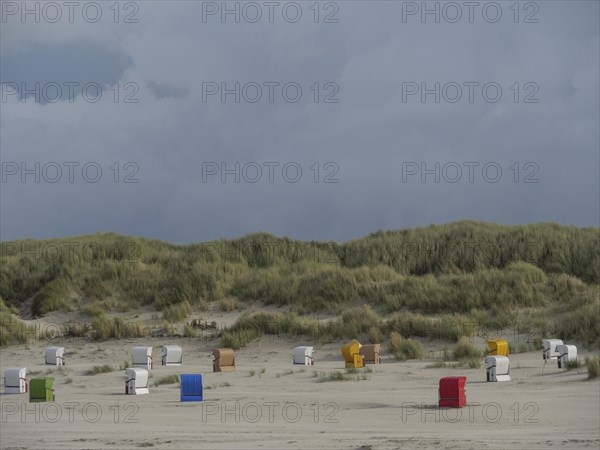 Beach chairs are spread out in front of a hilly dune landscape under a cloudy sky, colourful beach chairs on the beach and in the dunes under a cloudy sky, red wooden boat and yellow beach chair, Juist, Germany, Europe