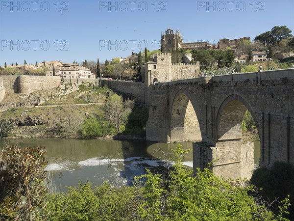Stone bridge with arches over a river, surrounded by a cityscape and vegetation, sunny weather, historic stone bridge with an old tower and an old town in the background over a wild river, Toledo, Spain, Europe