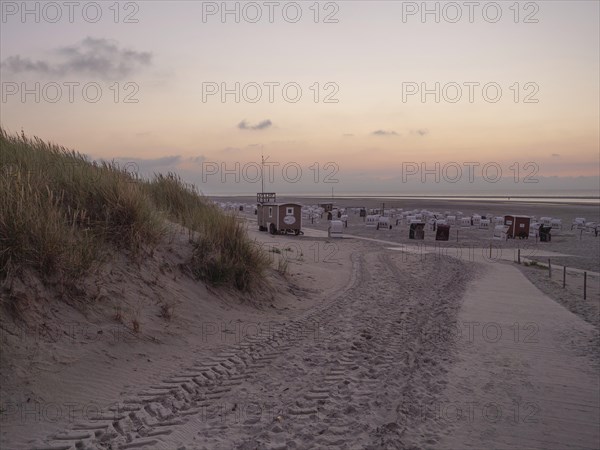 A peaceful path leads through the sand dunes to the beach during an orange sunset, setting sun on a beach with beach chairs and clouds in the sky, spiekeroog, germany