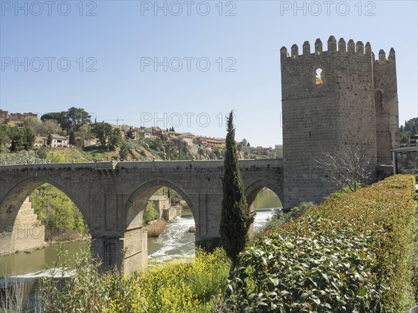 Stone bridge with arches and historic tower, surrounded by green landscape and river, sunny weather, historic stone bridge with an old tower and an old town in the background over a wild river, Toledo, Spain, Europe
