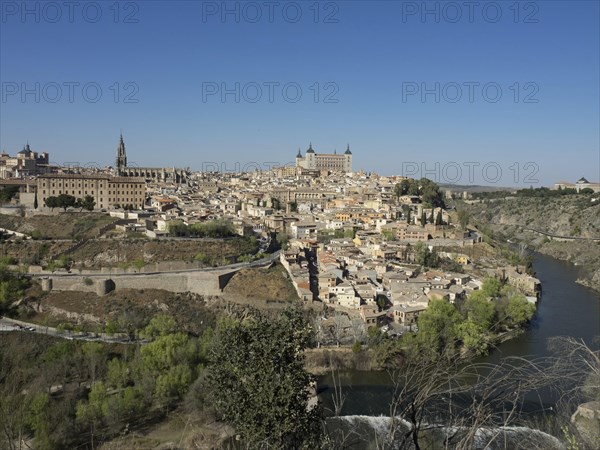 View of a historic riverside town with distinctive architecture and church towers, framed by hills and under a clear blue sky, historic town with old houses and large churches on a small river against a blue sky, toledo, spain