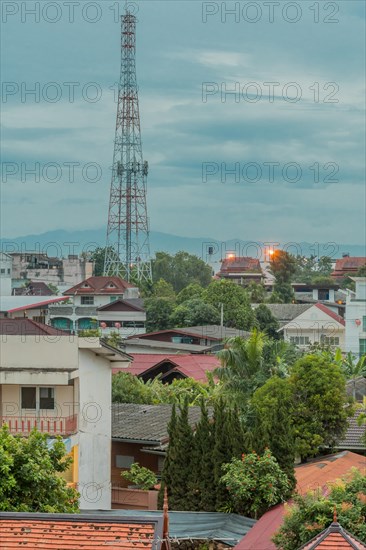 Communication tower above rooftops under cloudy morning sky. Taken from fourth story window in Thailand