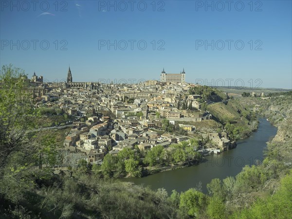 View of a historic riverside town with distinctive church towers and bridges, framed by hills under a clear blue sky, historic town with old houses and large churches on a small river against a blue sky, toledo, spain
