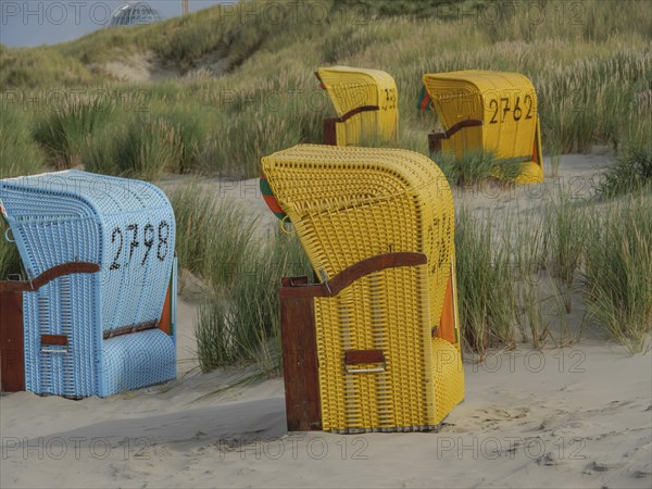 Colourful beach chairs standing on the sandy beach, surrounded by green dunes and a cloudy sky, colourful beach chairs on the beach and in the dunes under a cloudy sky, red wooden boat and yellow beach chair, Juist, Germany, Europe