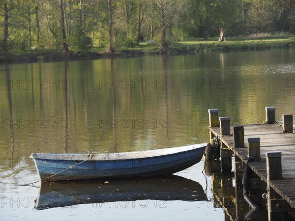 A boat is moored to a jetty and reflected in the calm waters of a lake, rowing boats and pedal boats on a small wooden jetty in the lake surrounded by trees, Borken Germany