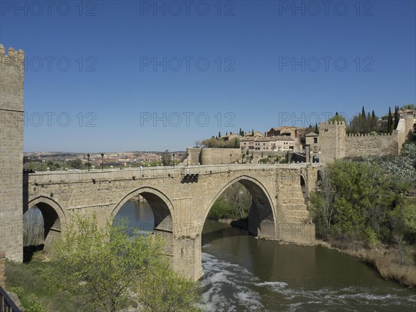 Historic stone bridge over a river surrounded by a city and nature, blue sky in the background, historic stone bridge with an old tower and an old city in the background over a wild river, Toledo, Spain, Europe