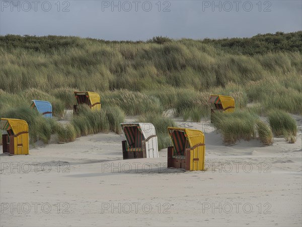 Several beach chairs scattered in front of high dunes under cloudy sky, colourful beach chairs on the beach and in the dunes under cloudy sky, red wooden boat and yellow beach chair, Juist, Germany, Europe