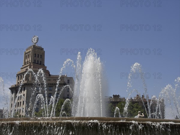 A fountain with powerful water jets in front of a historic building under a blue sky, fountain with figures in front of a historic building, barcelona, spain