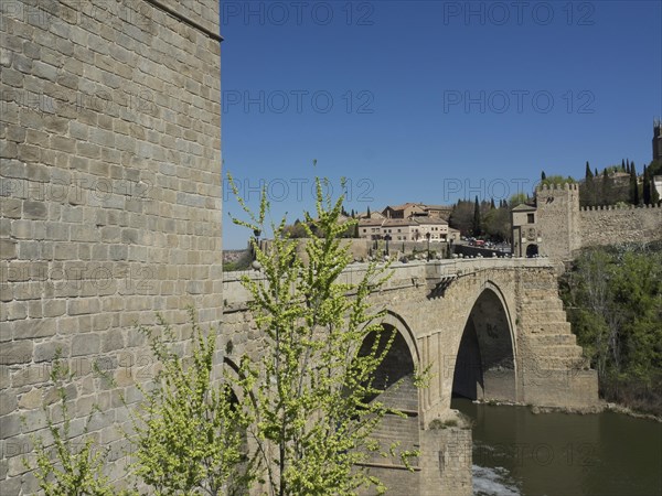 Historic stone bridge crossing a river, surrounded by green vegetation and clear blue sky, historic stone bridge with an old tower and an old town in the background over a wild river, Toledo, Spain, Europe