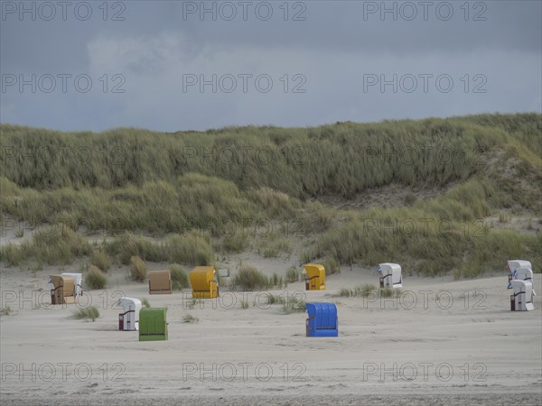 Different coloured beach chairs are spread out in front of a high dune landscape under a cloudy sky, colourful beach chairs on the beach and in the dunes under a cloudy sky, red wooden boat and yellow beach chair, Juist, Germany, Europe